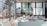 A spacious and modern cafeteria interior with multiple white tables and chairs. Large windows allow natural light to flood the area, providing a view of urban buildings and greenery outside. The setting is clean and organized, with wooden elements adding warmth to the space.