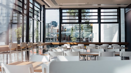 A spacious and modern cafeteria interior with multiple white tables and chairs. Large windows allow natural light to flood the area, providing a view of urban buildings and greenery outside. The setting is clean and organized, with wooden elements adding warmth to the space.