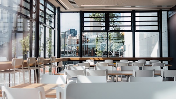 A spacious and modern cafeteria interior with multiple white tables and chairs. Large windows allow natural light to flood the area, providing a view of urban buildings and greenery outside. The setting is clean and organized, with wooden elements adding warmth to the space.