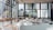 A spacious and modern cafeteria interior with multiple white tables and chairs. Large windows allow natural light to flood the area, providing a view of urban buildings and greenery outside. The setting is clean and organized, with wooden elements adding warmth to the space.