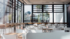 A spacious and modern cafeteria interior with multiple white tables and chairs. Large windows allow natural light to flood the area, providing a view of urban buildings and greenery outside. The setting is clean and organized, with wooden elements adding warmth to the space.