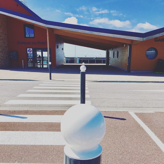 A modern building with a slanted roof and wooden exterior is located at a harbor. In front of the building, there is a crosswalk leading towards it. The building has a sign reading 'Bureau du Port.' The sky is clear with some clouds, suggesting a bright day.