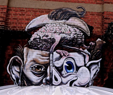 A surreal mural painted on a brick wall depicts a large human-like head split open at the top. A brain is visible, with a clawed hand reaching into it. The face displays distinctly different eyes, one human-like and the other cartoonish. The artwork combines realistic and abstract elements, merging the natural with the exaggerated.