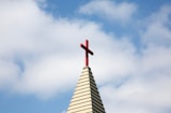 A church steeple with a red cross on top, set against a background of blue sky and white clouds.