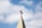 A church steeple with a red cross on top, set against a background of blue sky and white clouds.