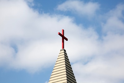A church steeple with a red cross on top, set against a background of blue sky and white clouds.