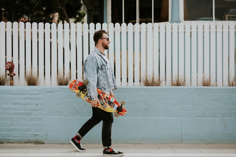 man carrying long board walking beside white fence