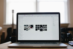 A laptop displaying a graphical design program with multiple small thumbnails on the screen, placed on a desk. Two camera lenses are positioned on either side of the laptop, and the background shows a softly lit room with a large window.