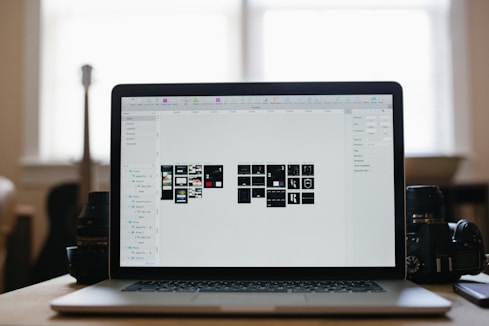 A laptop displaying a graphical design program with multiple small thumbnails on the screen, placed on a desk. Two camera lenses are positioned on either side of the laptop, and the background shows a softly lit room with a large window.