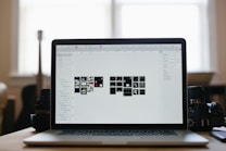 A laptop displaying a graphical design program with multiple small thumbnails on the screen, placed on a desk. Two camera lenses are positioned on either side of the laptop, and the background shows a softly lit room with a large window.