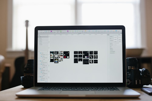 A laptop displaying a graphical design program with multiple small thumbnails on the screen, placed on a desk. Two camera lenses are positioned on either side of the laptop, and the background shows a softly lit room with a large window.