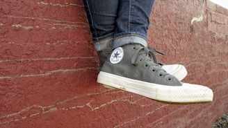 A pair of black and white high-top Converse sneakers rest against a textured, red brick wall. The person is wearing blue jeans, and the sneakers show signs of wear, adding a casual and laid-back feel.