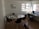 A small office space contains a white round table with several black chairs around it. On the table, there is a laptop, some papers, pens, and a microphone. On the side, a desk with a computer and a telephone is placed near a large window that lets in natural light. A diploma or certificate is framed on the wall. A dog is partially visible at the bottom of the image.