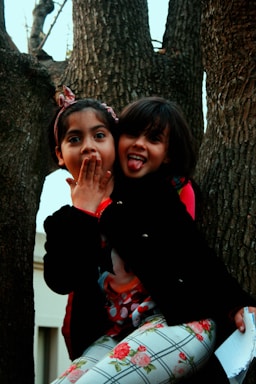 Two young children playfully pose while sitting between thick tree trunks. One child covers their mouth with surprise, while the other sticks out their tongue in a cheeky manner. Both are dressed in colorful clothing, and the natural setting provides a rustic backdrop.