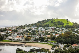 A scenic view of a suburban area featuring a green hill with a radio tower on top. The foreground includes a beach and waterfront area, while residential houses are scattered throughout, surrounded by lush vegetation. Cloudy skies hover above, creating a serene atmosphere.