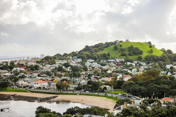 A scenic view of a suburban area featuring a green hill with a radio tower on top. The foreground includes a beach and waterfront area, while residential houses are scattered throughout, surrounded by lush vegetation. Cloudy skies hover above, creating a serene atmosphere.