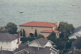 A house with a red-tiled roof is surrounded by trees and other buildings, overlooking a body of water. Two sailboats are visible on the water, suggesting a serene and picturesque coastal setting.