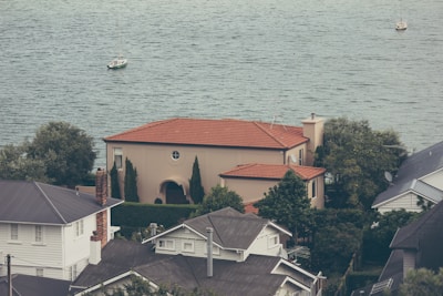 A house with a red-tiled roof is surrounded by trees and other buildings, overlooking a body of water. Two sailboats are visible on the water, suggesting a serene and picturesque coastal setting.
