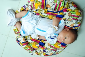 A baby is lying on a colorful, patterned cushion. The baby is wearing a white outfit with blue accents and white booties. The cushion features cartoon characters and vibrant patterns in colors such as yellow, pink, and blue.
