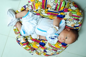 A baby is lying on a colorful, patterned cushion. The baby is wearing a white outfit with blue accents and white booties. The cushion features cartoon characters and vibrant patterns in colors such as yellow, pink, and blue.