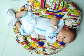A baby is lying on a colorful, patterned cushion. The baby is wearing a white outfit with blue accents and white booties. The cushion features cartoon characters and vibrant patterns in colors such as yellow, pink, and blue.
