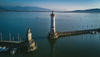 A lighthouse stands prominently on a pier extending into a large body of water. Nearby, a statue of a lion is mounted on a stone pedestal. Several flags line the pier, and boats are docked in the marina area. Mountains create a scenic backdrop under a clear blue sky.