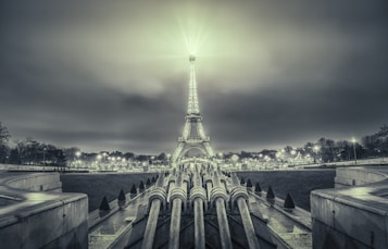 A monochrome image featuring the Eiffel Tower illuminated against a cloudy sky. The foreground consists of a series of fountains and geometrically aligned paths leading towards the tower. Surrounding the base of the tower, park lights and sparse trees are visible, creating a symmetrical composition.