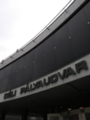 A large building facade with black panels and prominent block lettering spelling 'DÉLI PÁLYAUDVAR'. The structure suggests an entrance or front facing part of a larger complex, possibly a train station or public building. The weather appears overcast, contributing to a somewhat subdued atmosphere.