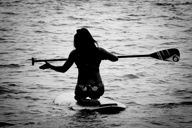 A person is kneeling on a paddleboard, holding a paddle horizontally over their shoulders. The scene is set on a body of water, with gentle waves surrounding the individual. The image is in black and white, creating a silhouette effect against the water.