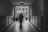 A black and white photograph featuring an elderly person walking through a dimly lit hallway towards a brighter outdoor area. The corridor has a tiled floor and wooden paneling on the walls. Outside, there is a courtyard with tables, chairs, and umbrellas, set against a backdrop of a brick building with arched windows.