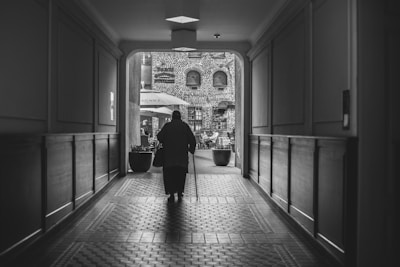 A black and white photograph featuring an elderly person walking through a dimly lit hallway towards a brighter outdoor area. The corridor has a tiled floor and wooden paneling on the walls. Outside, there is a courtyard with tables, chairs, and umbrellas, set against a backdrop of a brick building with arched windows.