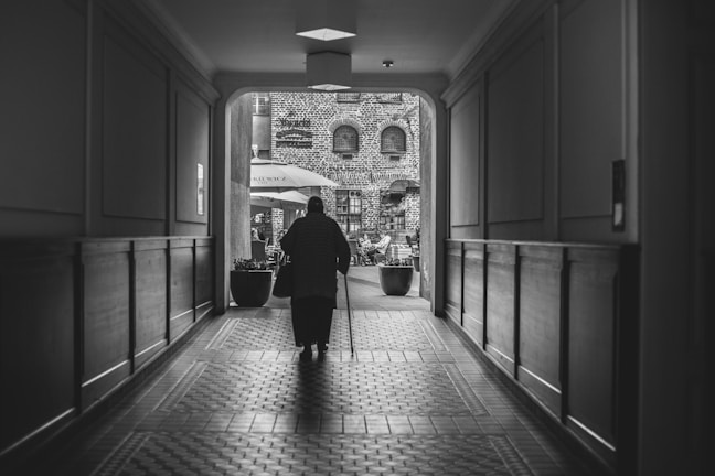 A black and white photograph featuring an elderly person walking through a dimly lit hallway towards a brighter outdoor area. The corridor has a tiled floor and wooden paneling on the walls. Outside, there is a courtyard with tables, chairs, and umbrellas, set against a backdrop of a brick building with arched windows.