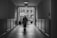 A black and white photograph featuring an elderly person walking through a dimly lit hallway towards a brighter outdoor area. The corridor has a tiled floor and wooden paneling on the walls. Outside, there is a courtyard with tables, chairs, and umbrellas, set against a backdrop of a brick building with arched windows.