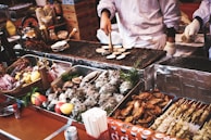 A bustling street food market scene featuring a vendor grilling a variety of seafood and meats. The table is filled with diverse shellfish, skewers, and garnishes such as lemons and tomatoes. The vendor, wearing gloves, is actively cooking on a wire mesh grill, surrounded by metallic trays displaying the fresh ingredients. The overall atmosphere is lively and inviting, with a focus on the enticing array of food offerings.