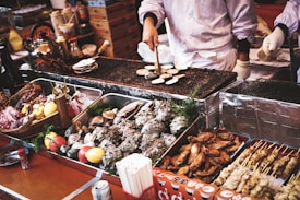 A bustling street food market scene featuring a vendor grilling a variety of seafood and meats. The table is filled with diverse shellfish, skewers, and garnishes such as lemons and tomatoes. The vendor, wearing gloves, is actively cooking on a wire mesh grill, surrounded by metallic trays displaying the fresh ingredients. The overall atmosphere is lively and inviting, with a focus on the enticing array of food offerings.