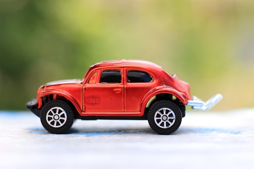 A red toy car with black windows and silver wheels is placed on a blue and white surface with a blurred green background.