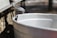 A metal spout is attached to a tree, with a small droplet of liquid poised at its tip, and an empty metal bucket is positioned beneath it to collect the liquid.