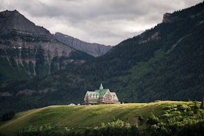 A picturesque lodge with green rooftops situated on a lush green hill, surrounded by towering forest-covered mountains under a cloudy sky.