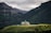 A picturesque lodge with green rooftops situated on a lush green hill, surrounded by towering forest-covered mountains under a cloudy sky.