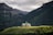 A picturesque lodge with green rooftops situated on a lush green hill, surrounded by towering forest-covered mountains under a cloudy sky.