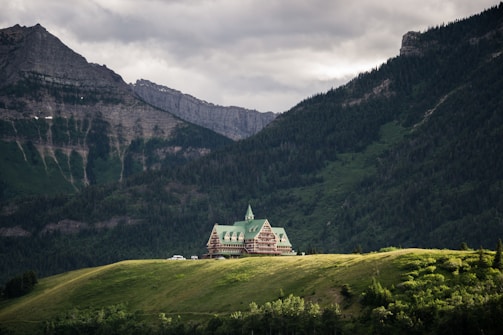 A picturesque lodge with green rooftops situated on a lush green hill, surrounded by towering forest-covered mountains under a cloudy sky.
