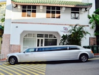 A long white limousine is parked on the street in front of a building with a green-tiled roof. The building has a sign that reads 'Louis Dreyfus Armateurs' and features a mix of brick and white plaster walls. Several green trash bins are visible on the right side of the image, alongside some palm trees and other plants.