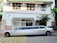 A long white limousine is parked on the street in front of a building with a green-tiled roof. The building has a sign that reads 'Louis Dreyfus Armateurs' and features a mix of brick and white plaster walls. Several green trash bins are visible on the right side of the image, alongside some palm trees and other plants.