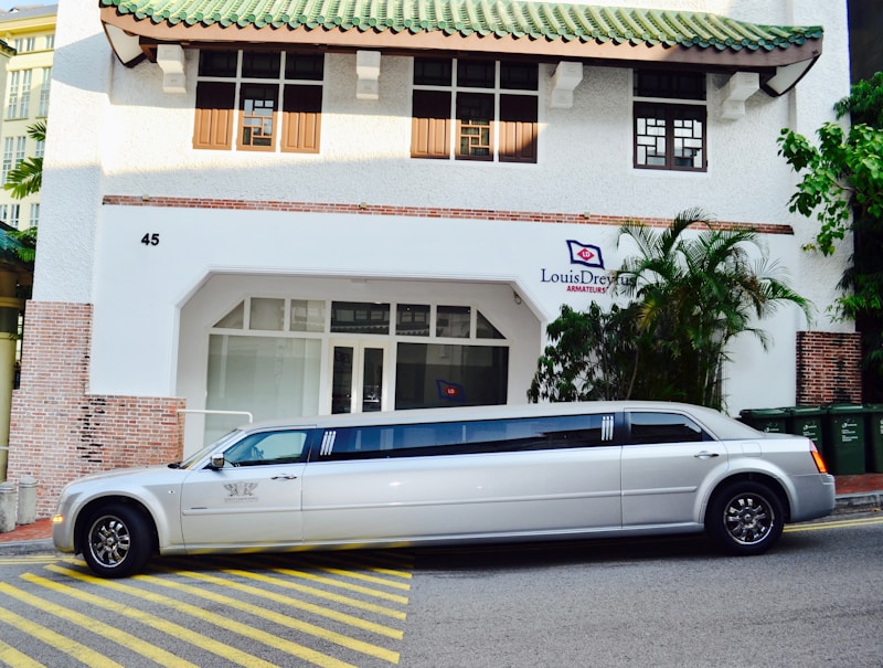 A long white limousine is parked on the street in front of a building with a green-tiled roof. The building has a sign that reads 'Louis Dreyfus Armateurs' and features a mix of brick and white plaster walls. Several green trash bins are visible on the right side of the image, alongside some palm trees and other plants.