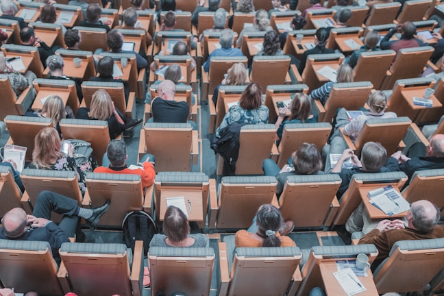 A large group of people seated in a lecture hall, viewed from behind. The attendees are sitting in wooden chairs with comfortable padding, arranged in orderly rows. Many individuals appear to be focused on reading materials or taking notes, with some holding pens and notebooks. A few cups and personal belongings are scattered among them, suggesting a long session.
