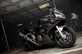 A black, sleek motorcycle is parked indoors against a chain-link fence. The design suggests a high-performance sports bike, with visible components such as the exhaust pipe, disc brakes, and curved bodywork. The lighting is low, creating contrasts that highlight the motorcycle's contours.