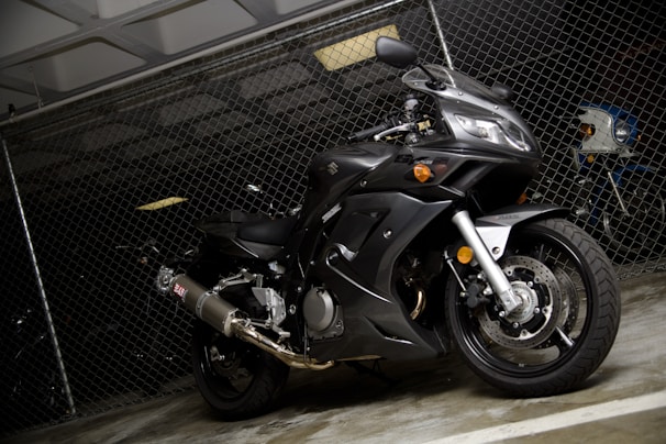 A black, sleek motorcycle is parked indoors against a chain-link fence. The design suggests a high-performance sports bike, with visible components such as the exhaust pipe, disc brakes, and curved bodywork. The lighting is low, creating contrasts that highlight the motorcycle's contours.
