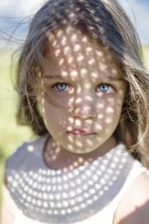 A young girl with blue eyes is looking directly at the camera. Her face is partially covered with a pattern of light and shadow, created by sunlight filtering through a mesh or lattice. The background is slightly blurred, emphasizing her facial features and the intricate play of light.