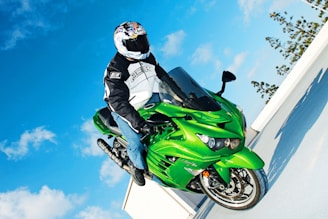 A person in a helmet and motorcycle gear is performing a wheelie on a vibrant green sportbike against a bright blue sky with scattered clouds. The bike and rider appear dynamic and in motion, showing a stunt performed on a rooftop or elevated area, with trees visible in the background.