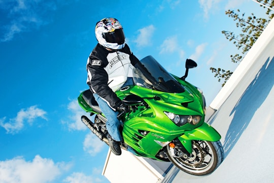 A person in a helmet and motorcycle gear is performing a wheelie on a vibrant green sportbike against a bright blue sky with scattered clouds. The bike and rider appear dynamic and in motion, showing a stunt performed on a rooftop or elevated area, with trees visible in the background.